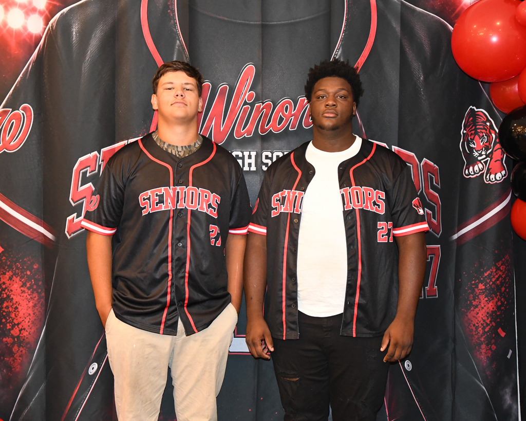 Two individuals stand next to each other in front of a backdrop with a baseball jersey design. One wears a black jersey, the other a white shirt and black pants.