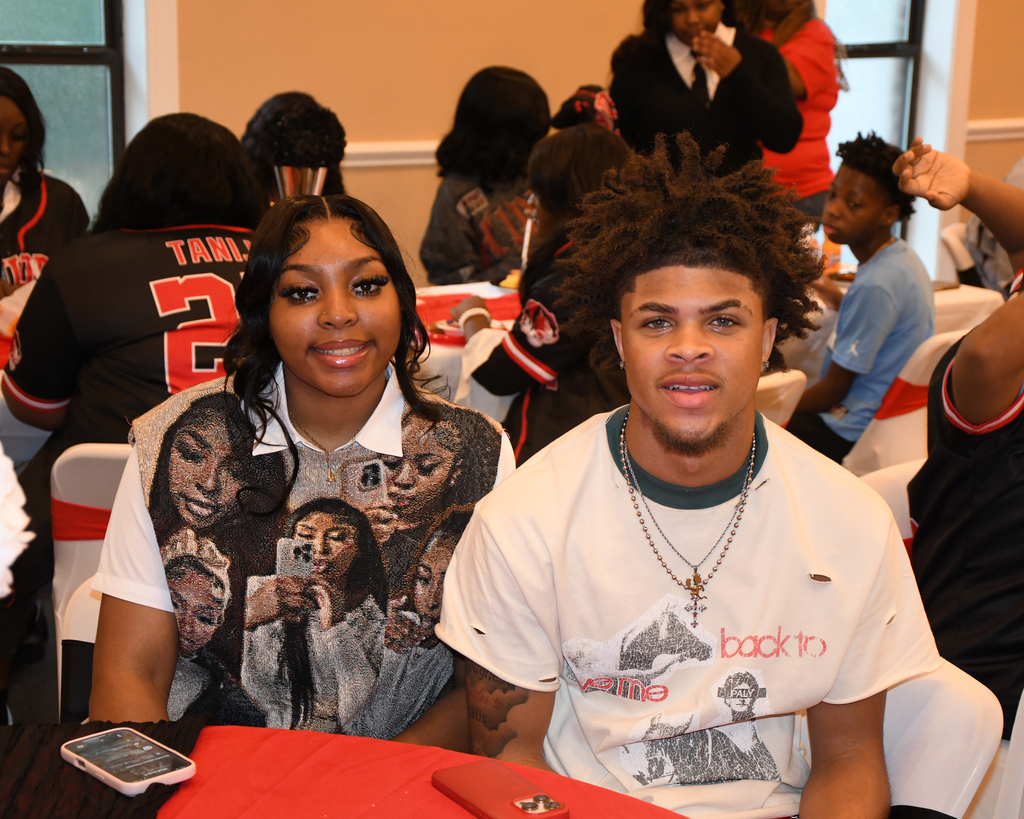 A woman and man sitting at a table with others, the woman wearing a shirt with pictures of women.