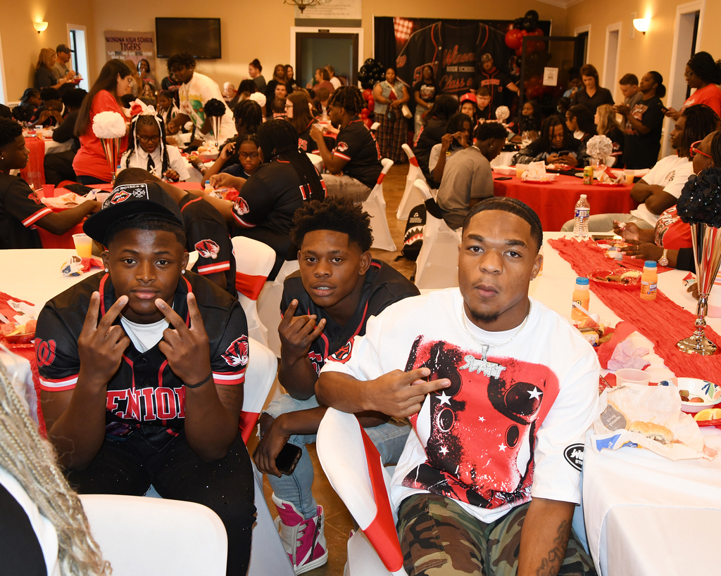 Many people seated at tables, some wearing matching jerseys. Two men in front wearing jerseys, one making a hand sign.