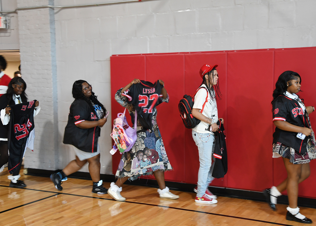 Five individuals walk together in a gymnasium. Four wear jerseys with the number 23. One person wears a red cap.