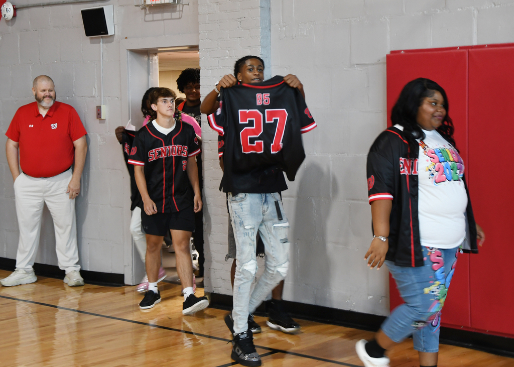 Four people on a basketball court; a man holds a jersey with the number 27.