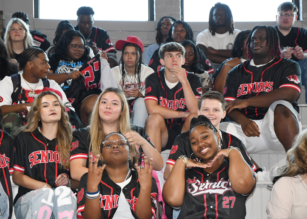 A crowd of students, some in red and black jerseys, are seated in bleachers, some are smiling and interacting.
