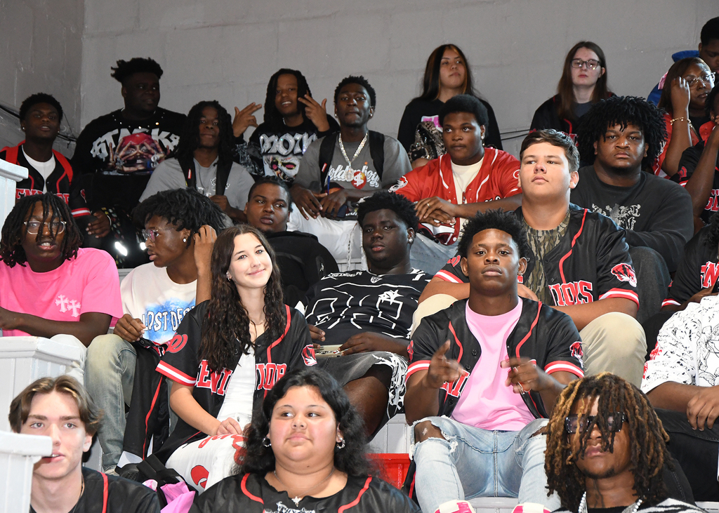 A group of students in varied attires seated in a school gym, some wearing sports jerseys.
