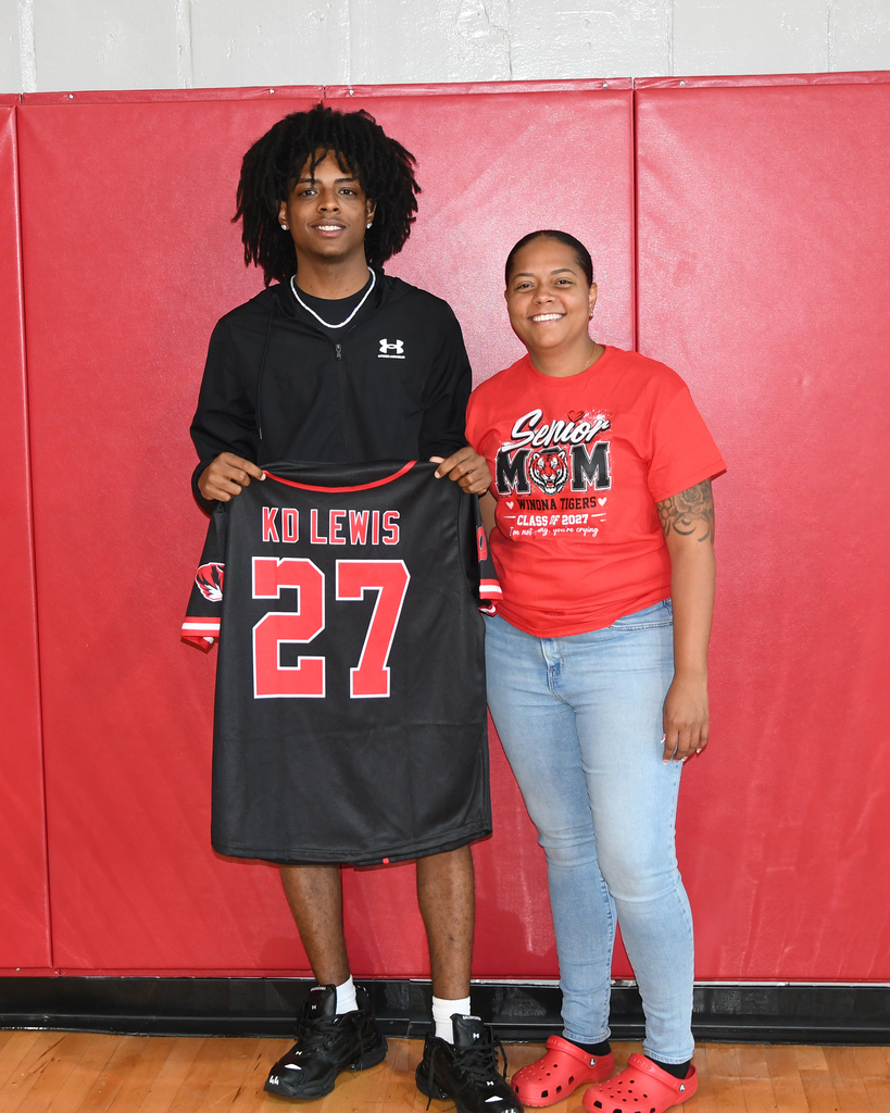 Two individuals stand together, one holding a black jersey with red numbers. The jersey says "KD LEWIS 27." They are in front of a red wall.