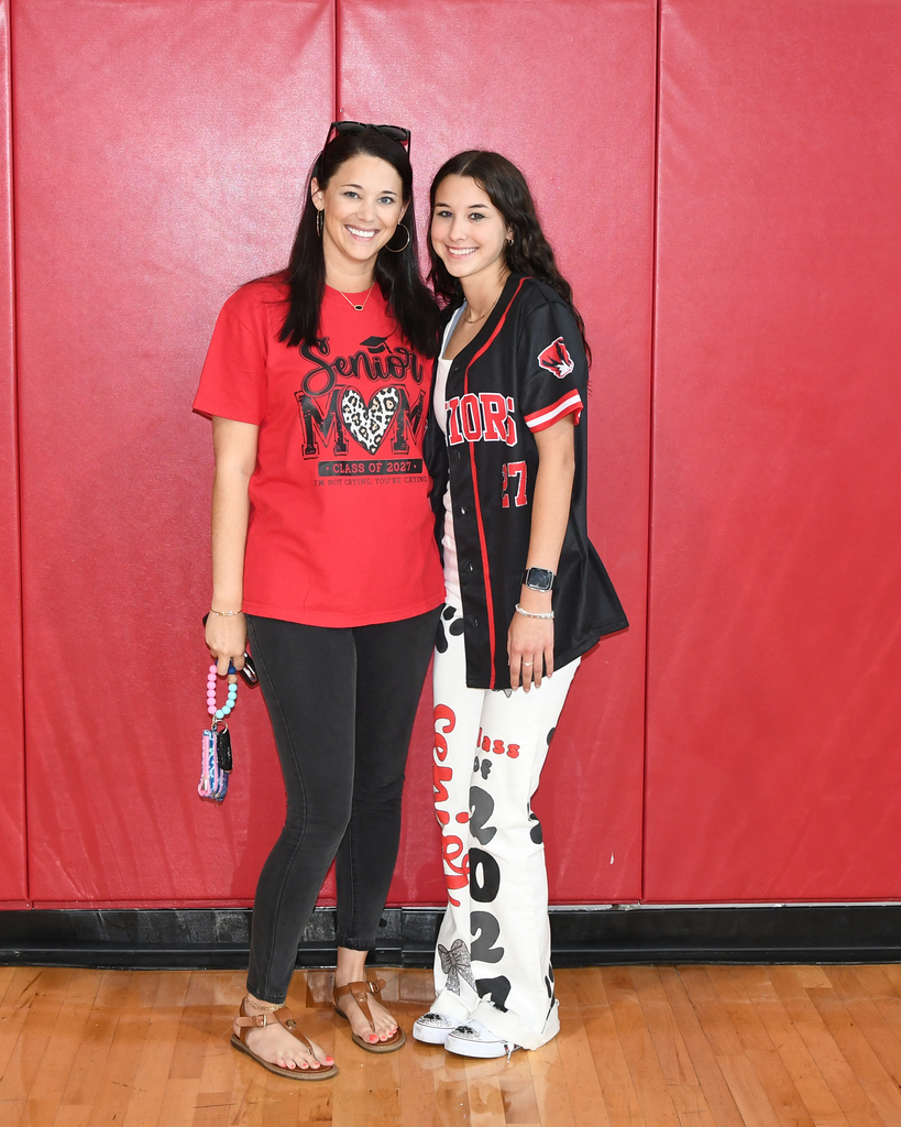 Two women in sports jerseys stand side by side in front of a red wall.