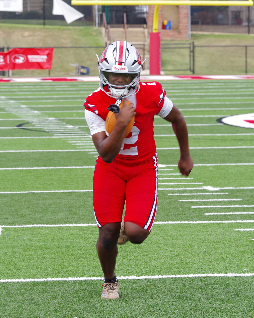 A football player in red, white, and silver uniform, helmet, and shoes runs with a football on a field.