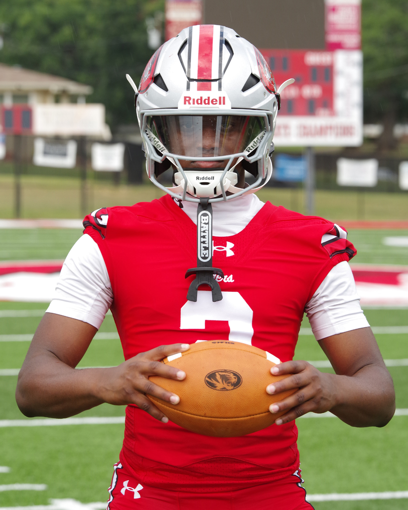 A football player in a red jersey, helmet with "Riddell" logo, holding a ball on a grassy field.