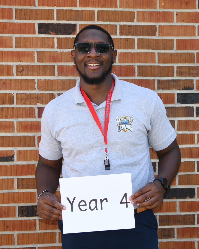 A man stands in front of a brick wall holding a sign that reads "Year 4." He wears sunglasses and a gray polo with a red lanyard.
