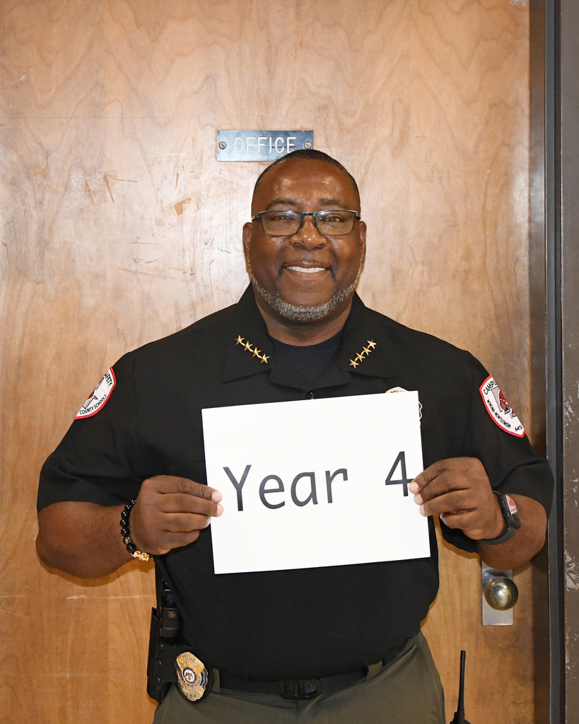 A person in a police uniform holds a sign with "Year 4" against a wooden door.