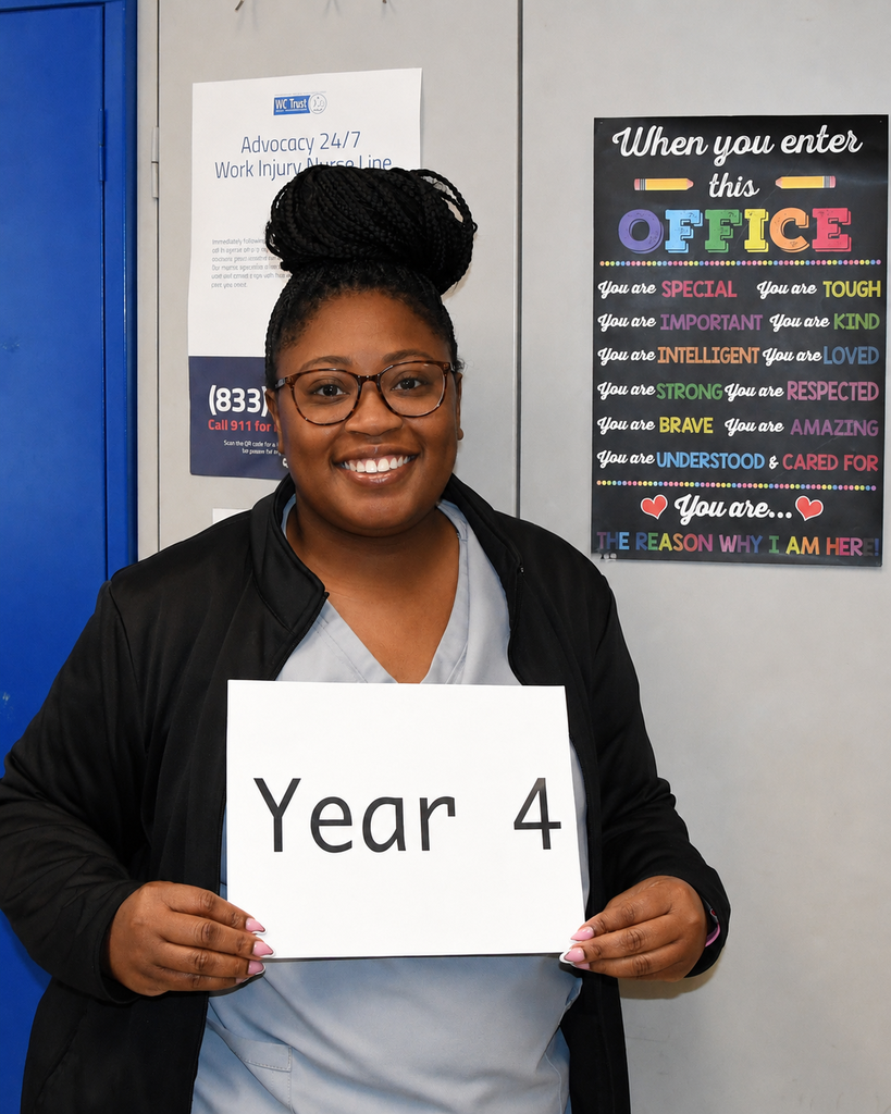 A smiling woman holds a sign that reads "Year 4" in front of a white wall with motivational posters.