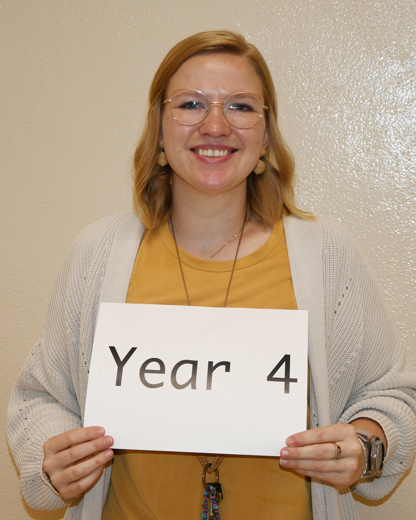 A woman in a yellow shirt, gray cardigan, and eyeglasses holds a white sign that reads "Year 4."