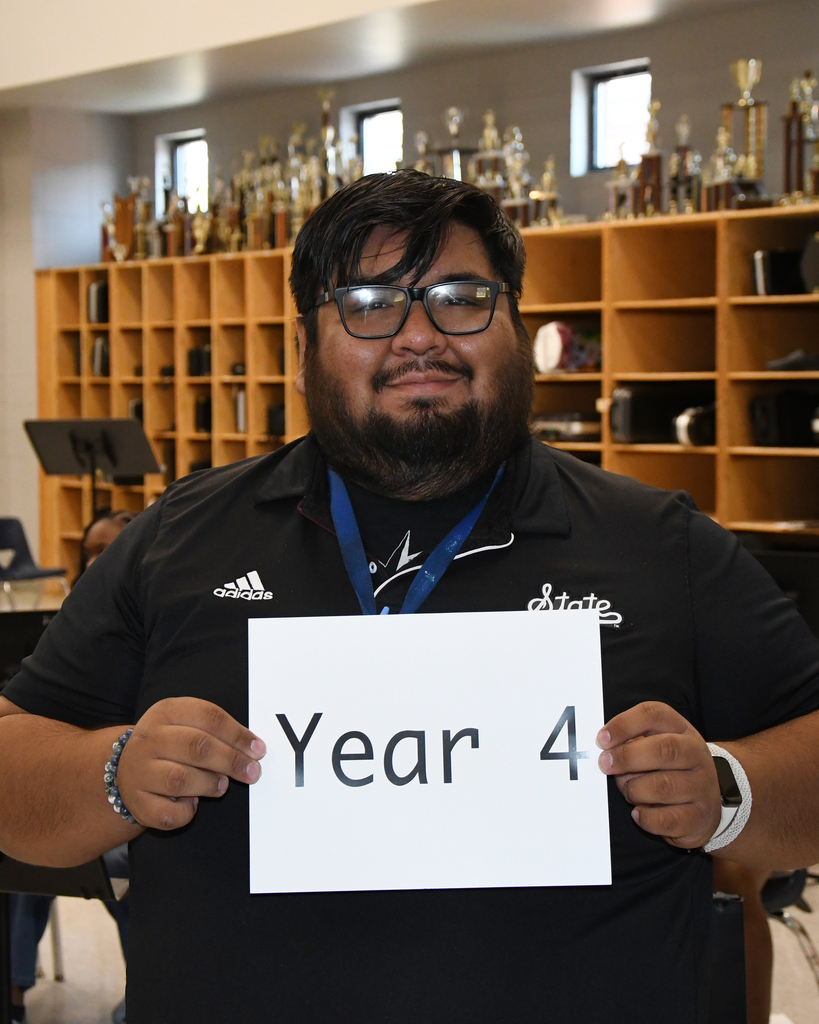Person holding a white card with "Year 4" in front of shelves with trophies and a music stand.