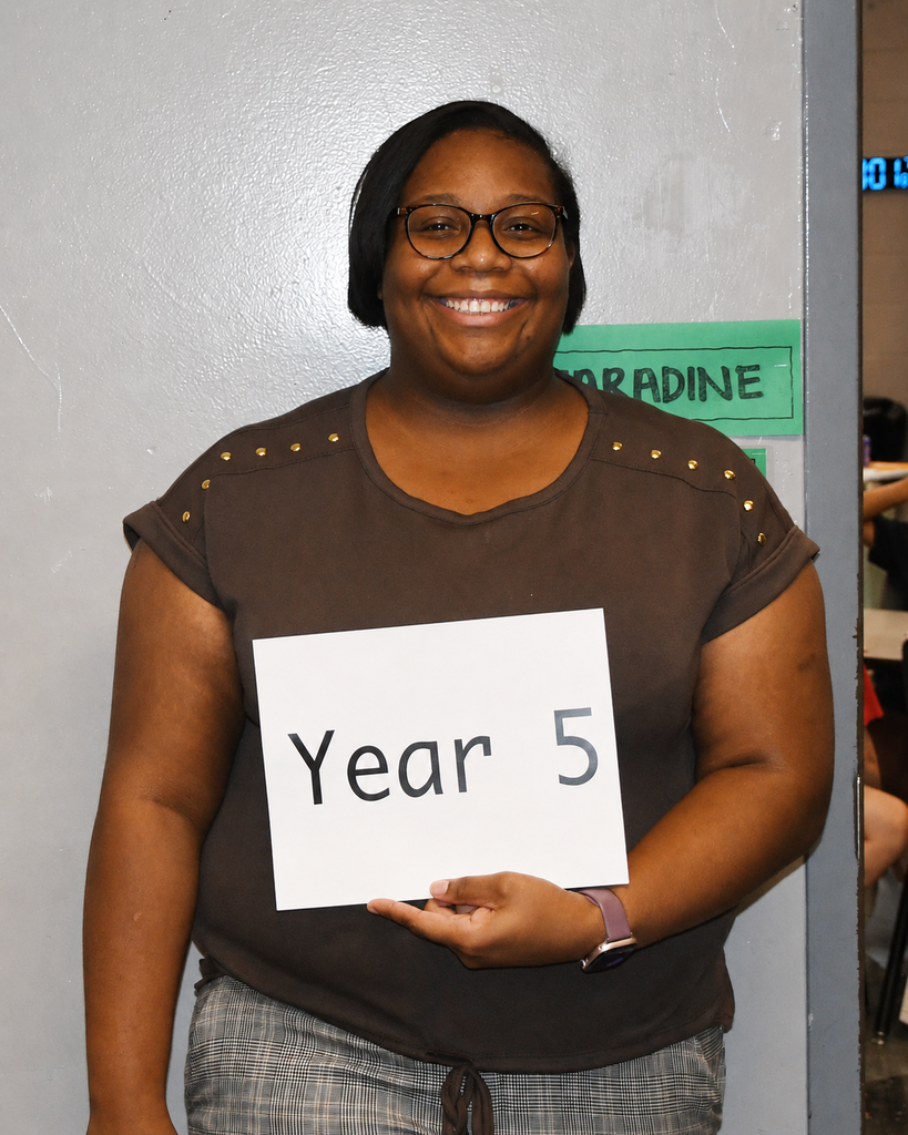 A woman smiling, holding a white sign that reads "Year 5." She is in front of a light-colored wall.
