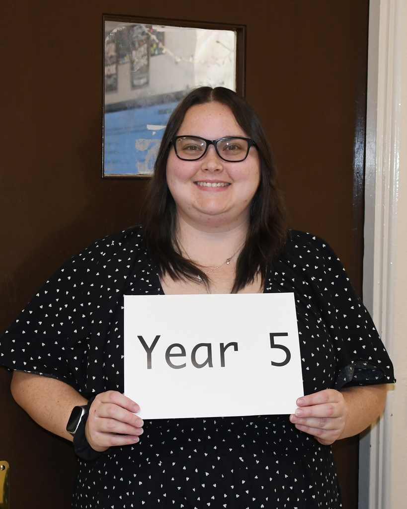 A smiling person holding a white sign that reads "Year 5" stands in front of a brown door.