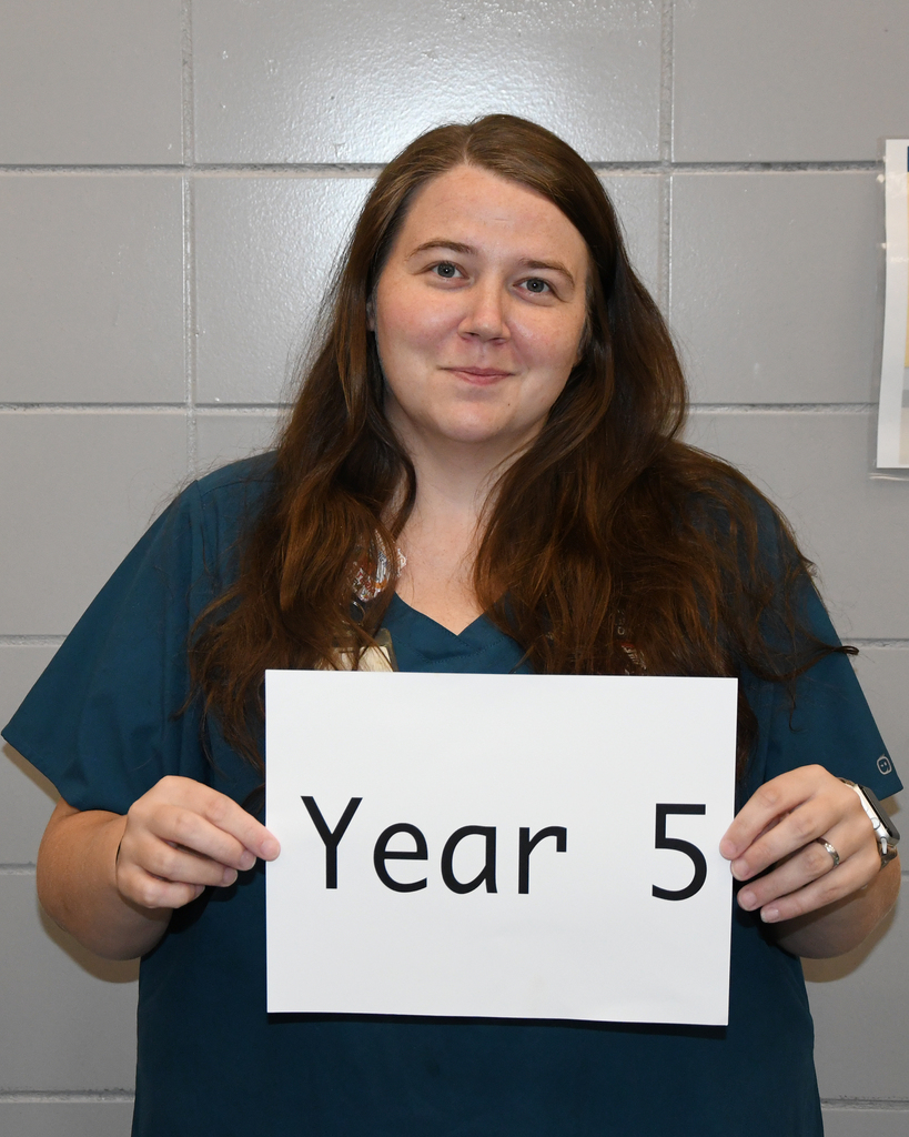 Person in blue scrubs holds a sign with "Year 5" on it, standing before a gray wall.