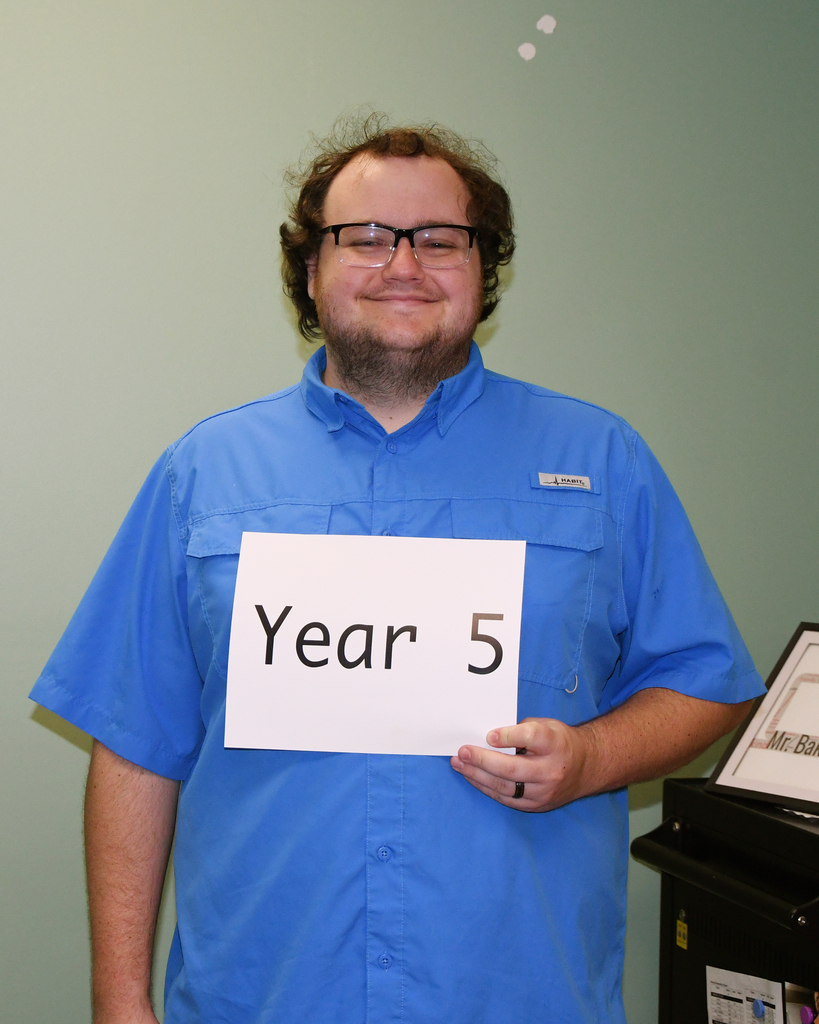 A person in a blue shirt holds a white sign with black text reading "Year 5". They have a smile on their face and are standing against a green wall.