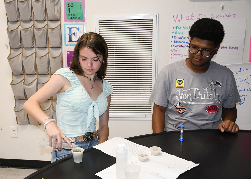 Two students, a girl in a light blue top and a boy in a gray t-shirt, conduct an experiment in a classroom.
