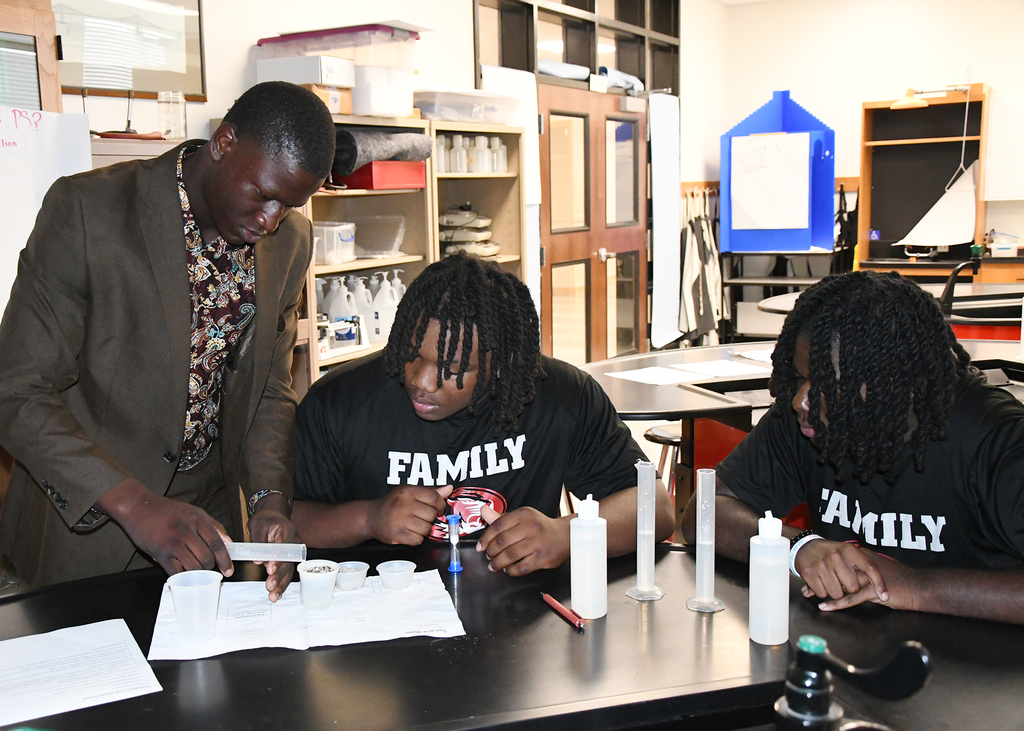 Three people in a classroom with a teacher, focusing on a chemistry experiment with lab equipment.