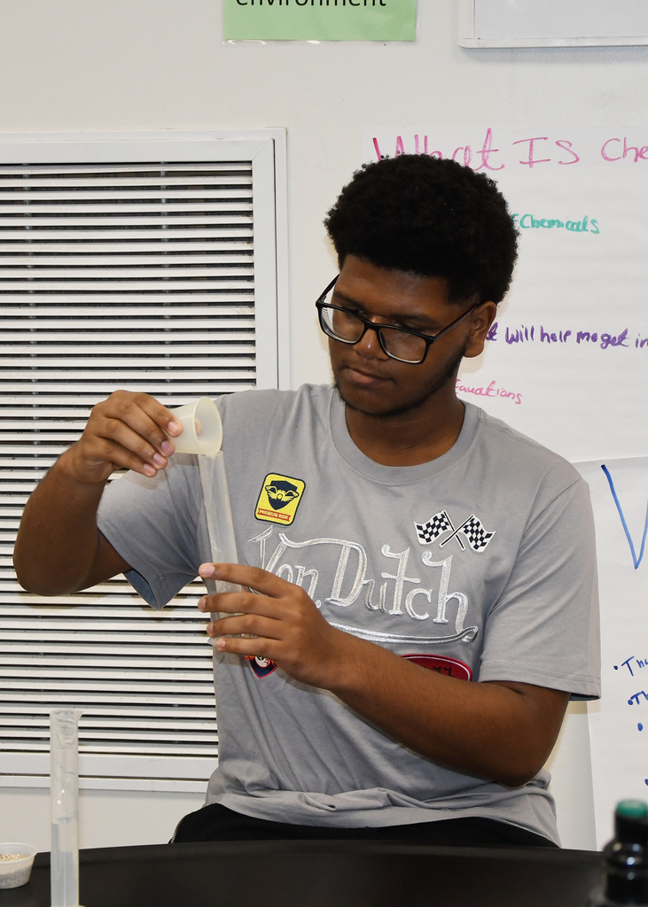 Person with glasses in a lab, holding a transparent tube, seated at a table. Behind them, a whiteboard with writing.