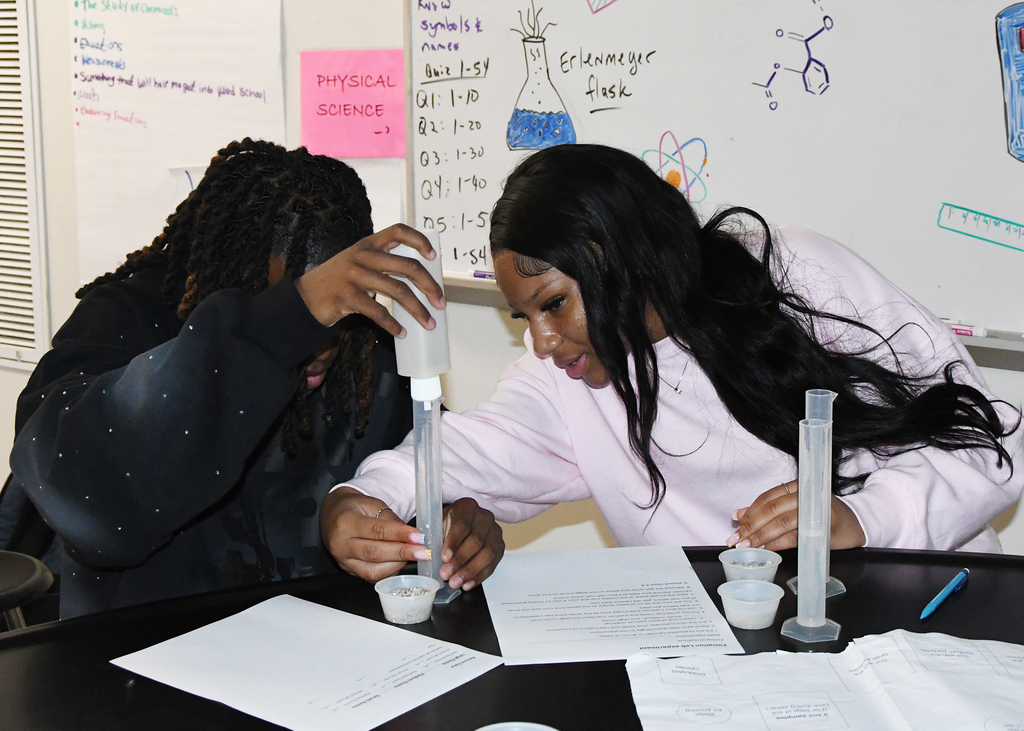 Two students working at a table, one pouring liquid into a test tube, other holding a paper.