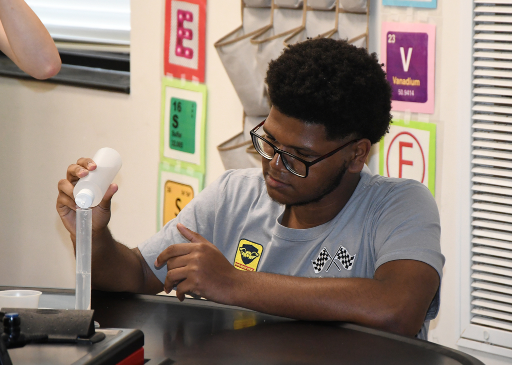 A person wearing glasses sits at a desk, pouring a liquid from a white bottle.