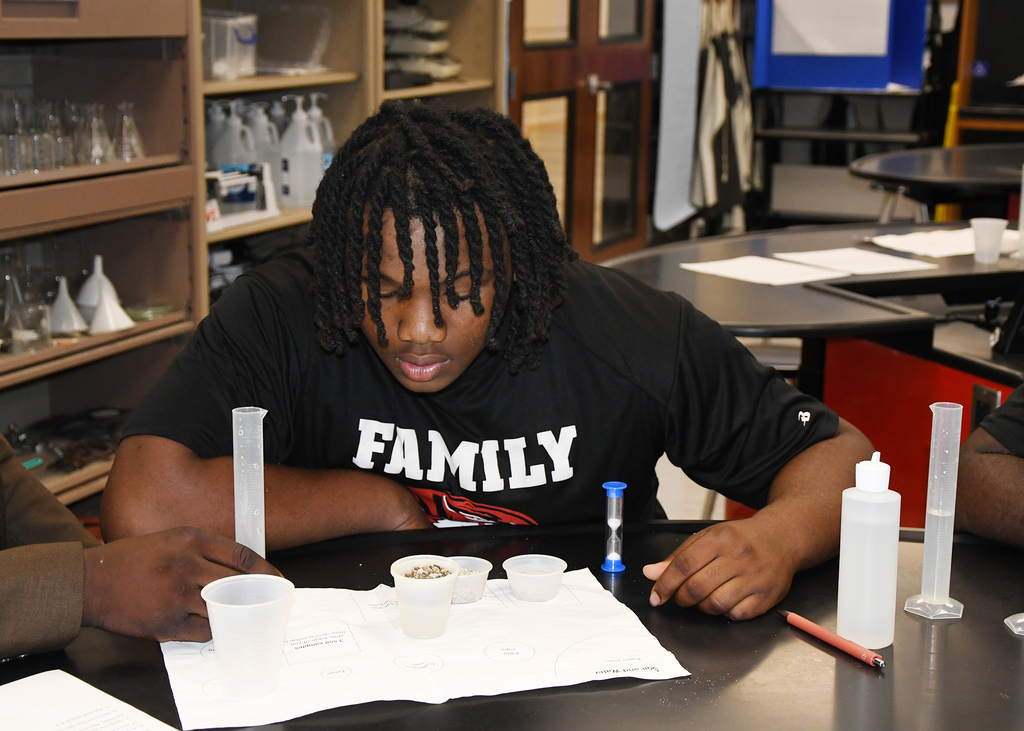 A young man sits at a table, looking down, with beakers, measuring cups, and other lab equipment.