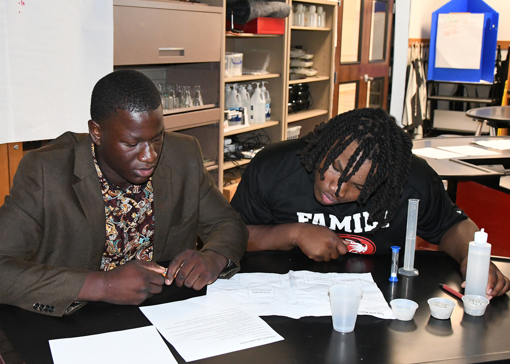 Two people work together at a table with papers, cups, and lab equipment. One wears a gray suit, and the other wears a black shirt.