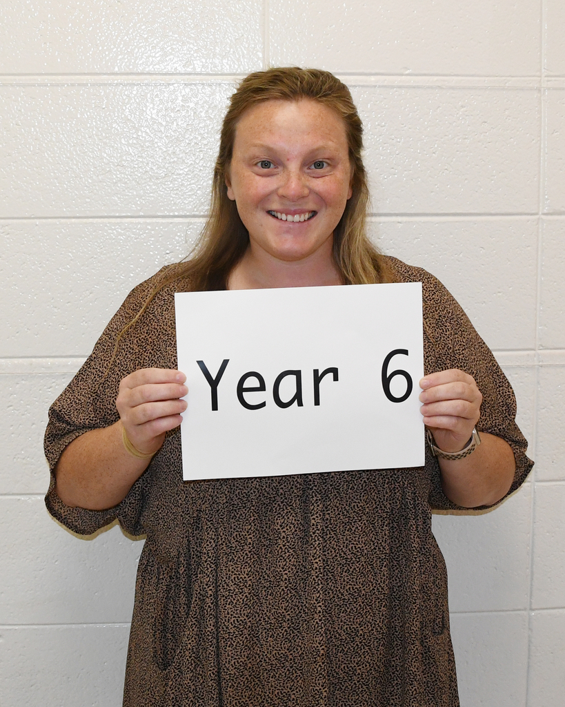 Woman holding a white sign with the text "Year 6" in black letters against a white wall.