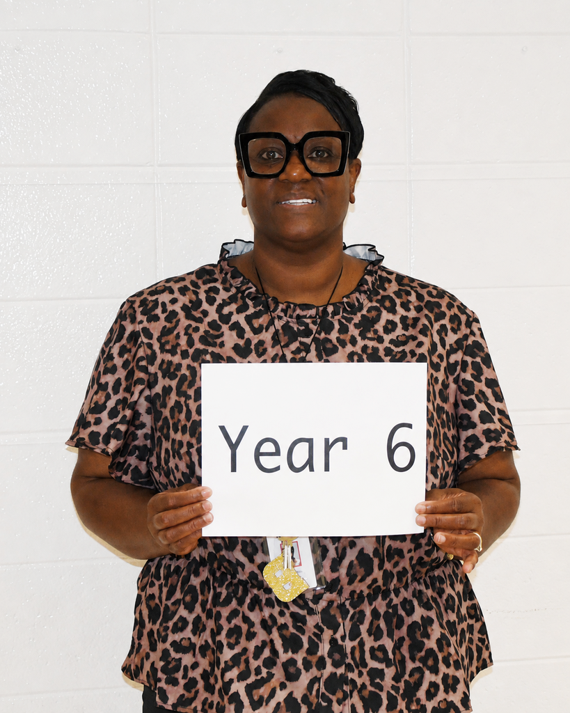 Woman wearing glasses and a leopard print blouse holds a sign reading "Year 6" against a white background.