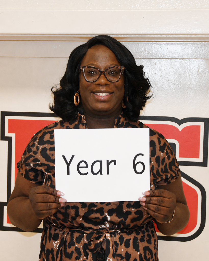 Woman in a leopard print dress and glasses holds a sign with "Year 6" in front of a red and white logo.