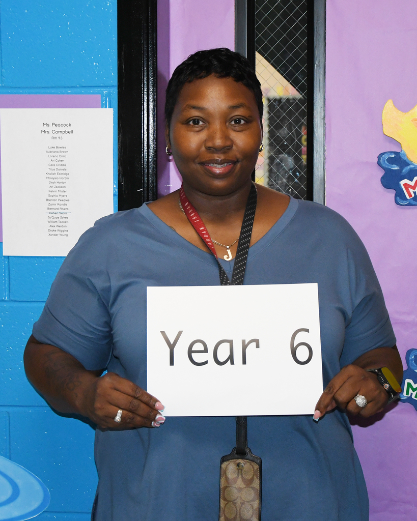 A woman in blue stands with a white sign reading "Year 6" in front of a colorful classroom wall.