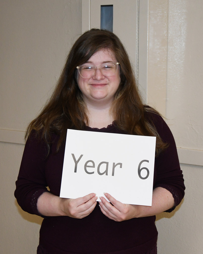 A woman in glasses holds a white sign with black letters reading "Year 6." She is wearing a maroon top.