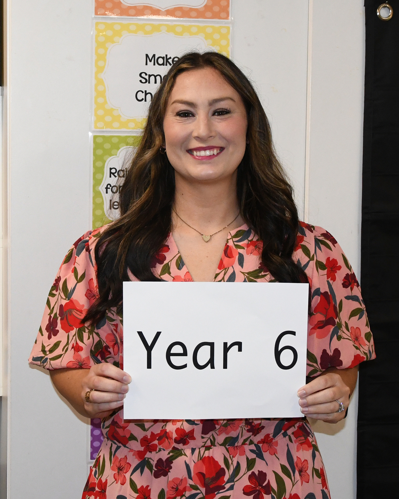 A smiling woman holding a sign that reads "Year 6". She wears a pink floral dress and a necklace.