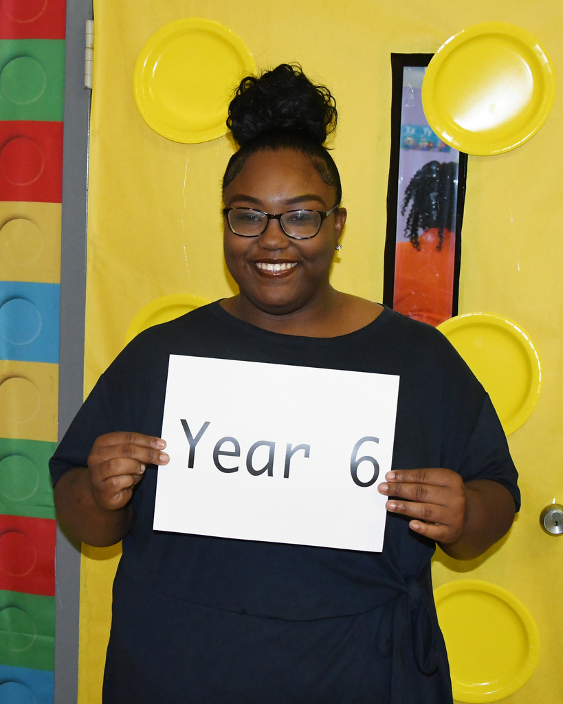Person holding sign reading "Year 6" in front of a yellow door with yellow plates and colorful blocks.