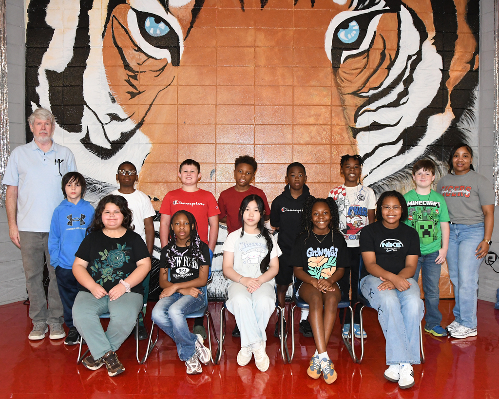 A group of young people sits in chairs before a tiger mural on a wall. One adult stands behind them.
