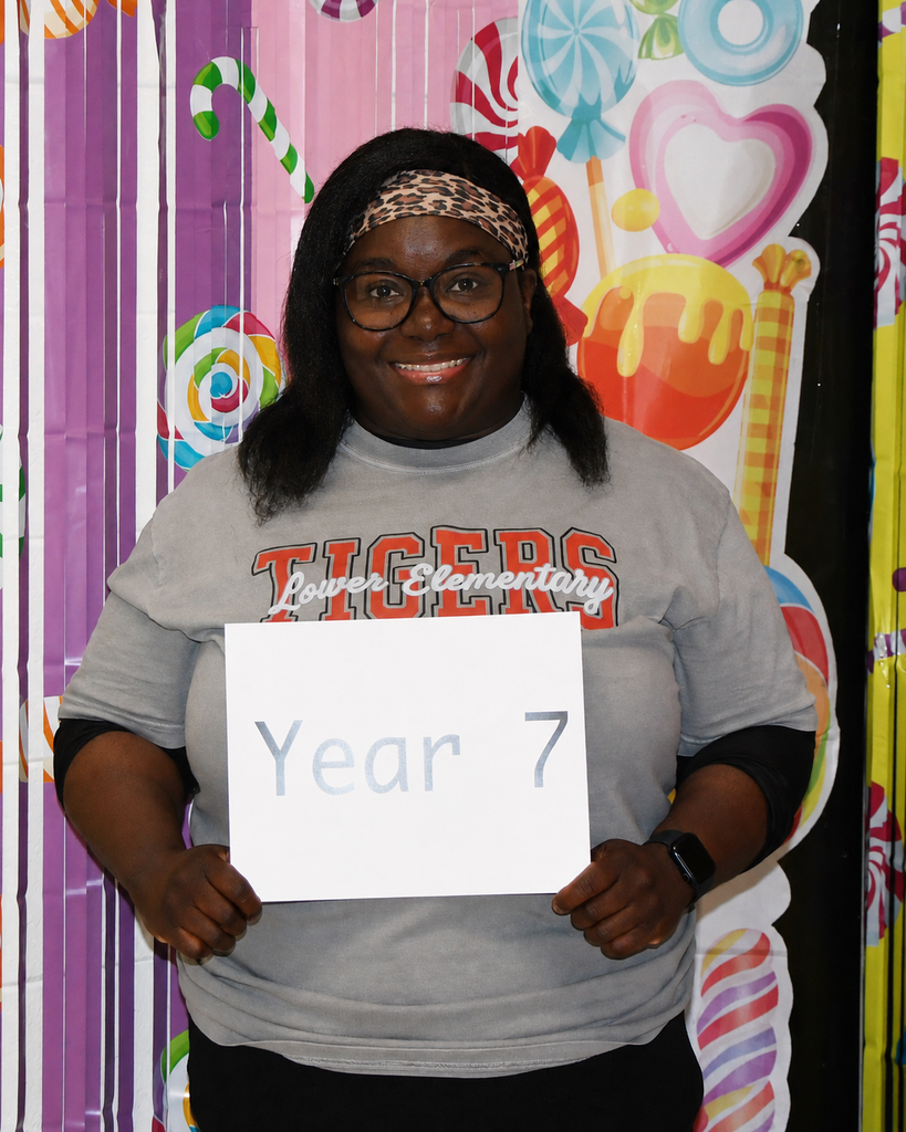 Person in glasses holds sign reading "Year 7" against backdrop of candy designs.