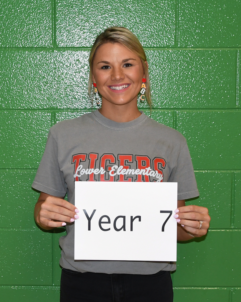 A smiling individual holds a white sign with black text that says "Year 7" in front of a green wall.