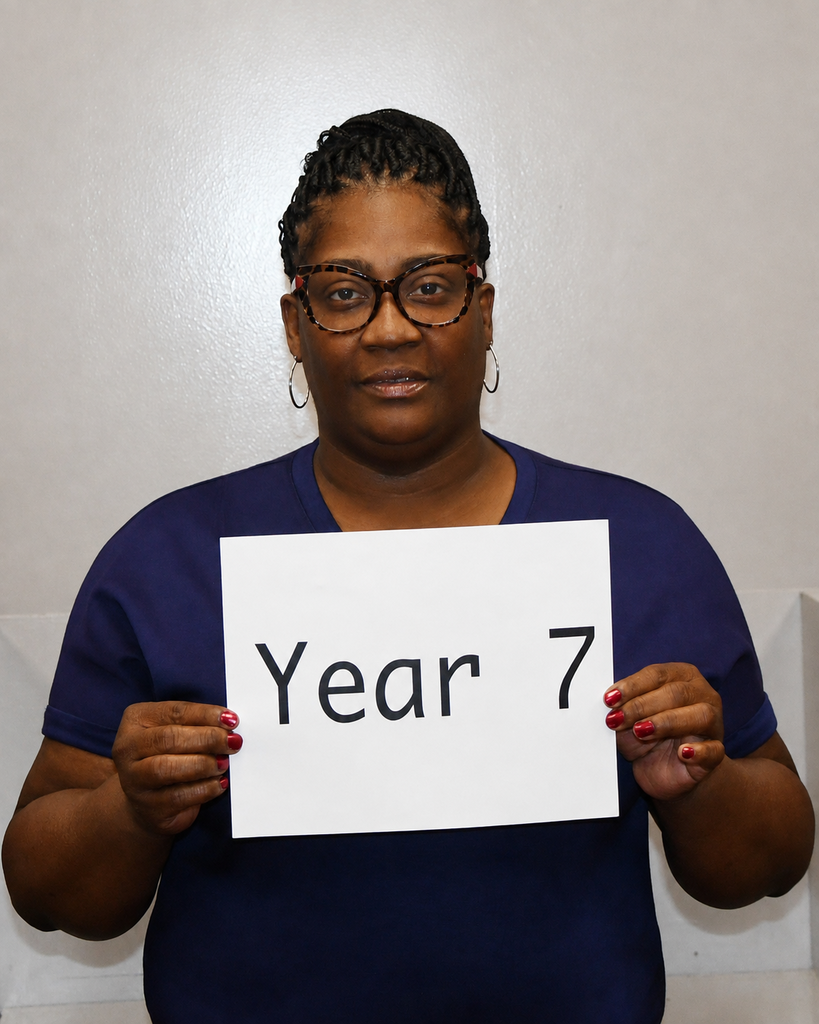 A person in a blue shirt and glasses holds a white sign that reads "Year 7" in front of a white wall.