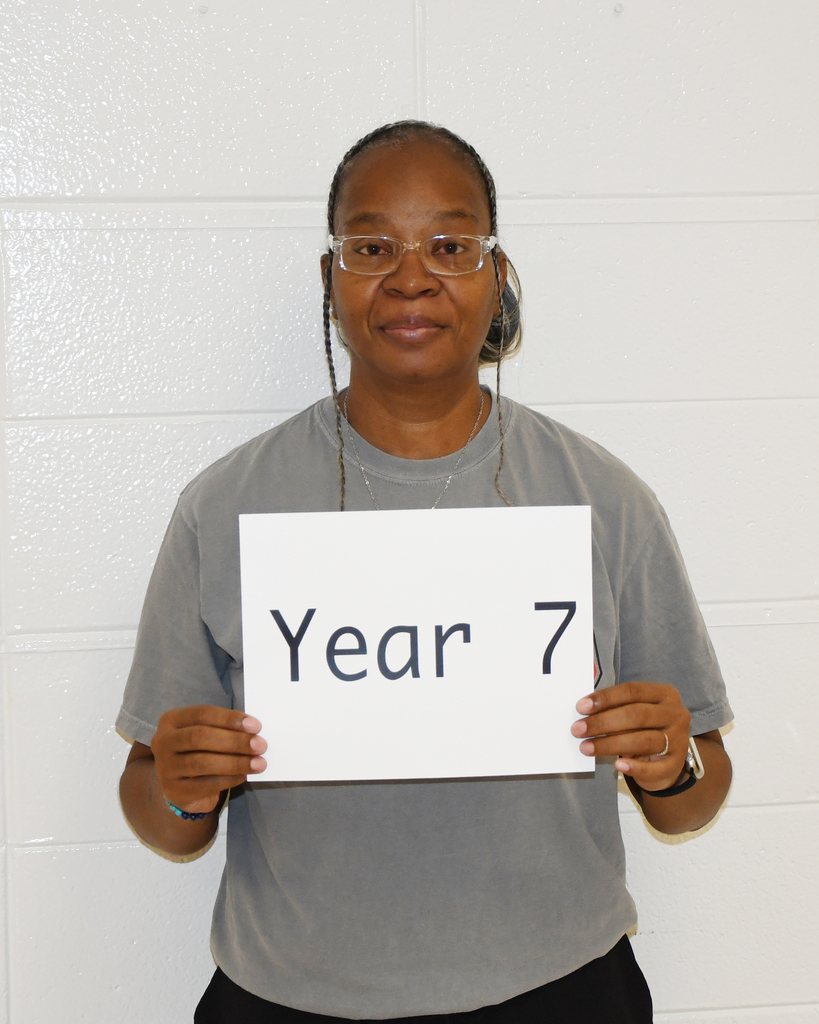 Person in gray shirt holds a white sign with "Year 7" on it against a white wall.