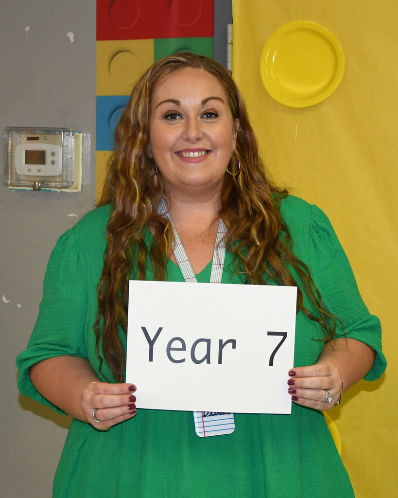 Woman in green dress smiling and holding a sign that says "Year 7" in front of a yellow wall.