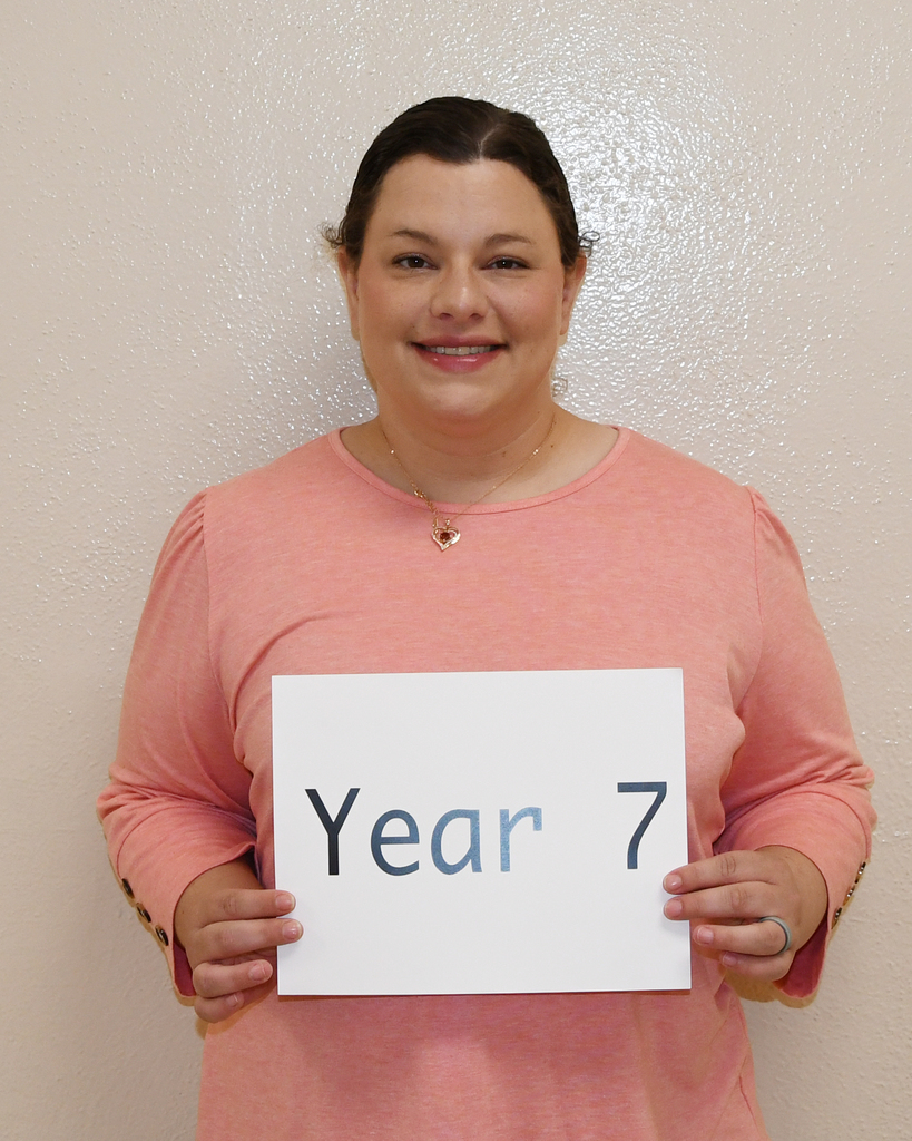 A person in a pink shirt holding a white sign that reads "Year 7." They are smiling.