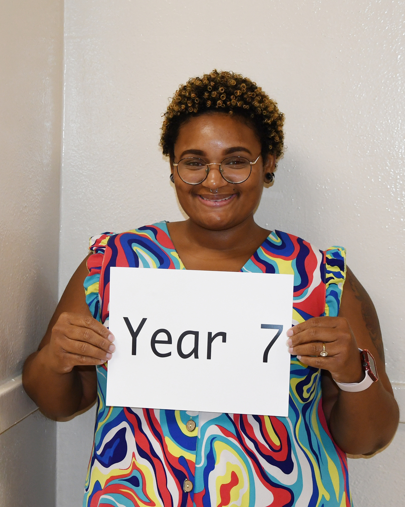 A woman with short hair, glasses, and a colorful top smiles while holding a sign saying "Year 7".