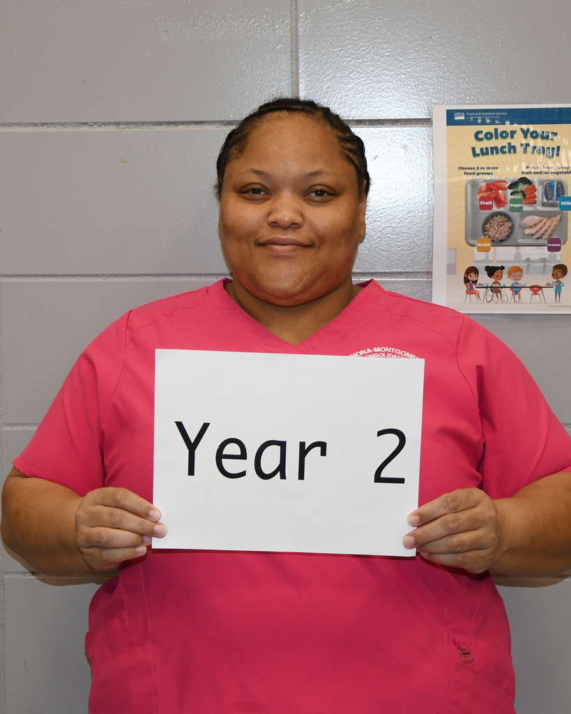 Person in pink scrubs holding a white sign with "Year 2" in front of a gray wall with a colorful poster.
