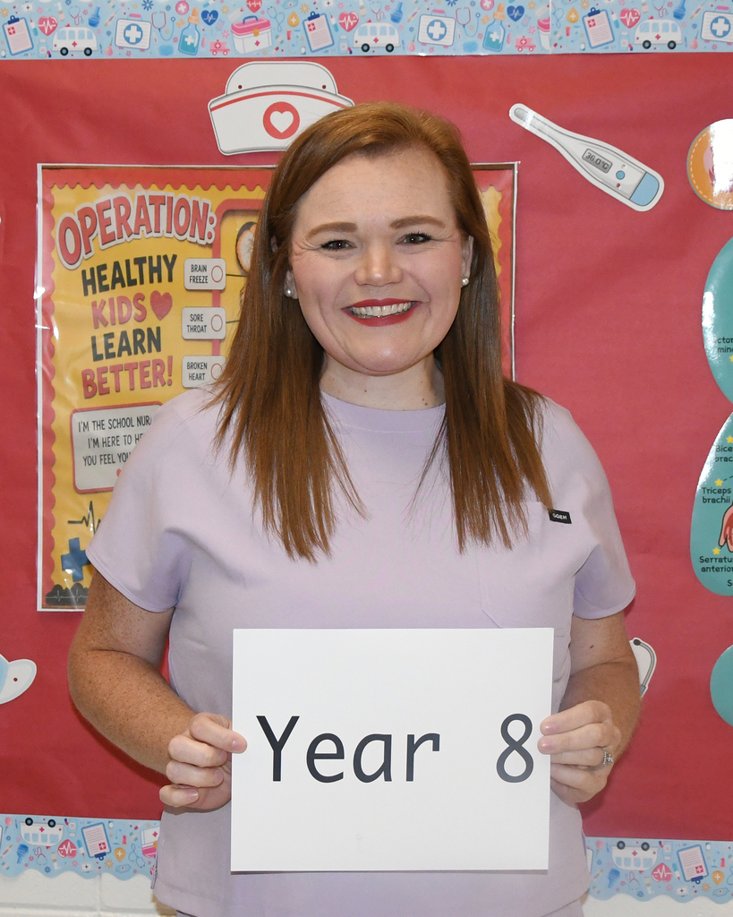 Woman holding a sign that says "Year 8" in front of a red backdrop with medical posters.