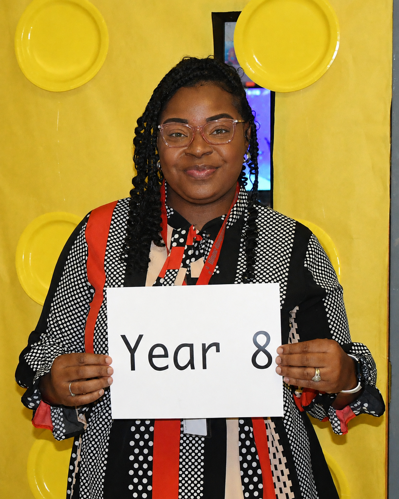 Person holding a white sign with black text "Year 8" in front of a yellow wall with large circular cutouts.