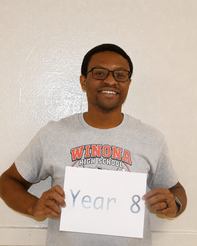 A smiling man in a gray Winona High School shirt holds a white card with blue text reading "Year 8."