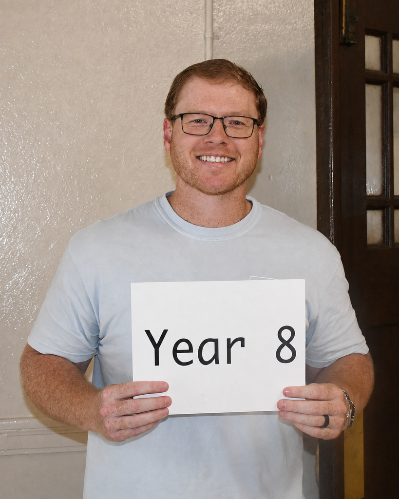 A person in a light blue shirt holds a white sign with "Year 8" in front of a beige wall and open door.