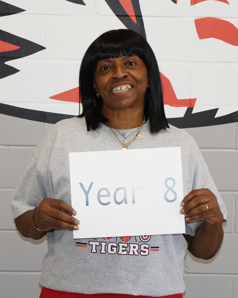 A woman holds a sign reading "Year 8" in front of a wall with a sports team logo.