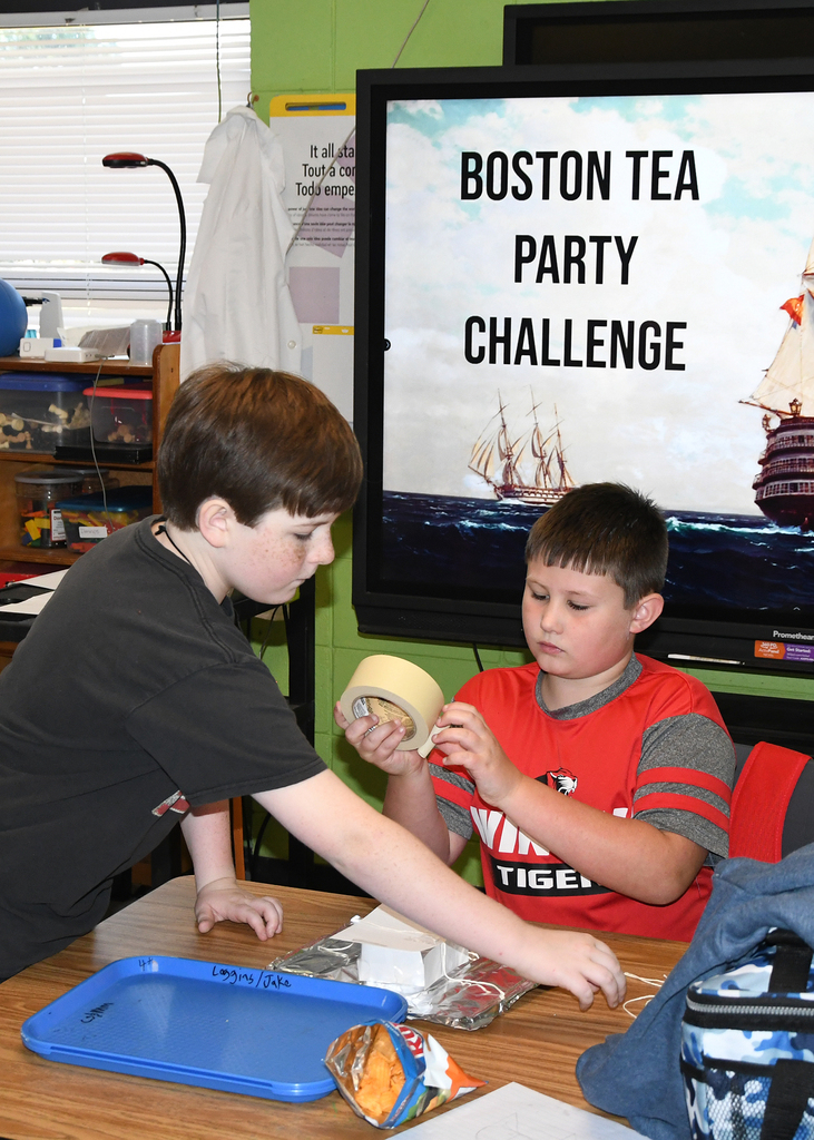 Two boys at a table, one taping, the other watching. A screen in the background displays "Boston Tea Party Challenge."