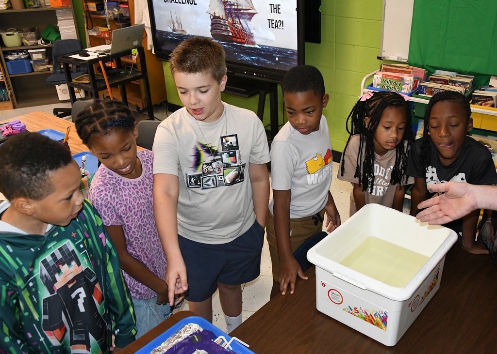 Several children gather around a table in a classroom, looking at a white container filled with water.
